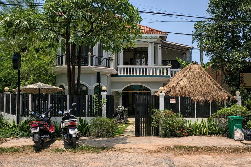 The white two-storey villa by day behind its dark wooden fence and thatched sala